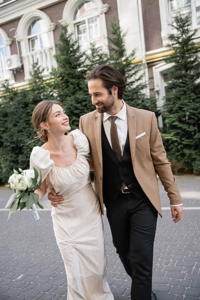 Happy young bride in white dress holding wedding bouquet while walking with bearded groom on street — Foto stock