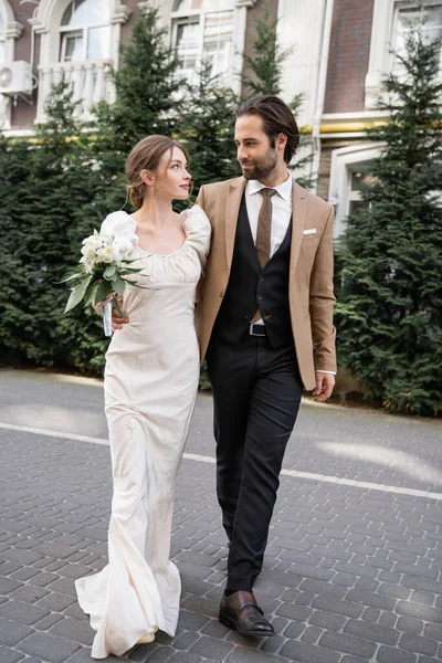 Full length of gorgeous bride in white dress holding wedding bouquet while standing with bearded groom on street — Stock Photo
