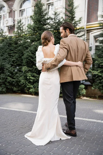 Back view of bride in white dress hugging with bearded groom on street — Photo de stock
