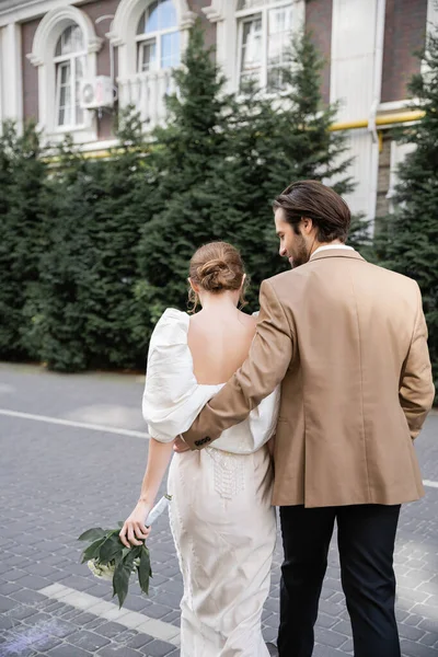 Back view of bride in white dress holding wedding bouquet and walking with bearded groom on street — Stockfoto