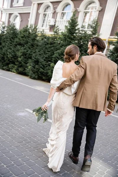 Back view of bride in white dress holding wedding bouquet and walking with groom on street — Stockfoto
