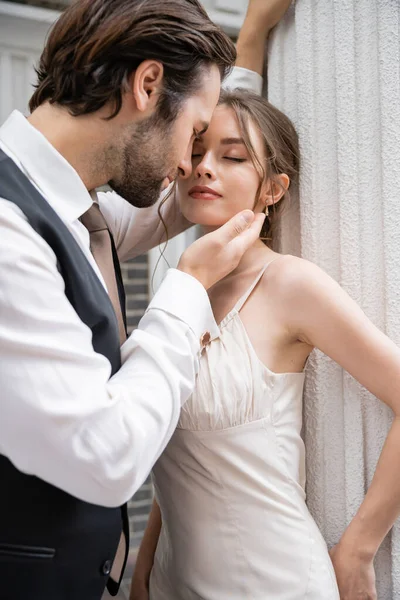 Bearded groom in formal wear touching face of pretty bride with closed eyes - foto de stock