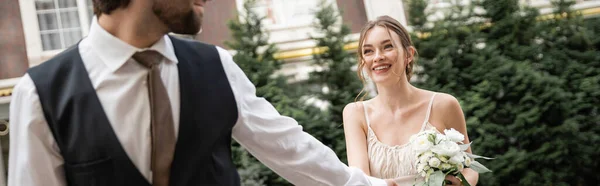 Happy bride in white dress with wedding bouquet holding hands with groom while walking outside, banner — Foto stock