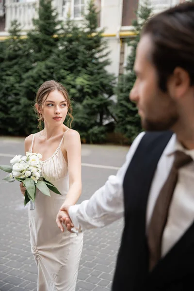 Novio borroso en chaleco de la mano de la novia hermosa en vestido blanco con ramo de boda - foto de stock