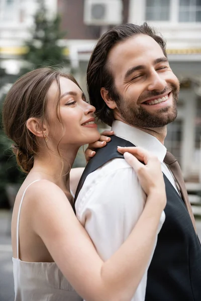 Mujer joven en vestido de novia abrazando novio feliz en chaleco - foto de stock