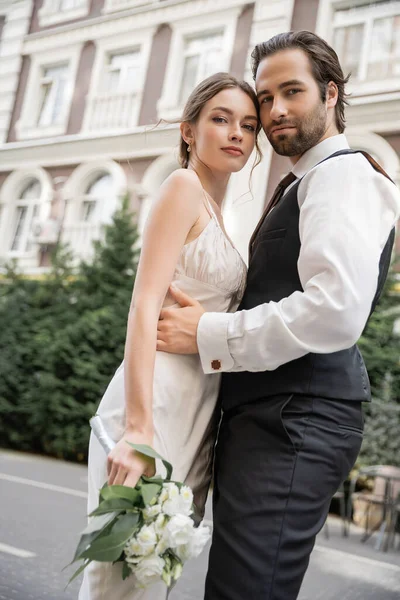 Low angle view of bearded groom in vest embracing bride in white dress with wedding bouquet — Stock Photo