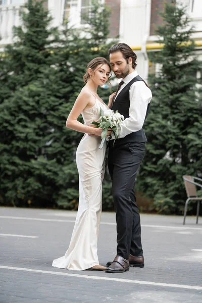 Full length of bearded man in vest embracing bride in white dress with wedding bouquet — Foto stock