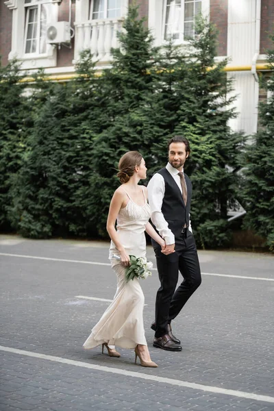 Tutta la lunghezza di uomo in vestito che tiene la mano di sposa in vestito bianco con mazzo di fiori — Foto stock