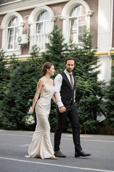 Full length of groom walking with happy bride in white dress holding bouquet of flowers — Photo de stock