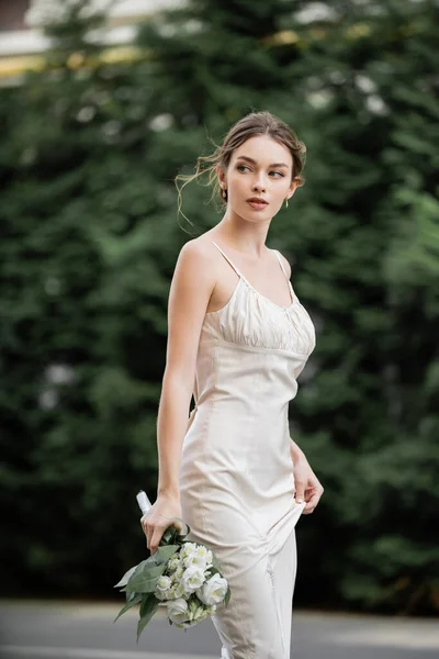 Giovane donna in abito da sposa in possesso di bouquet con fiori in fiore e guardando fuori — Foto stock