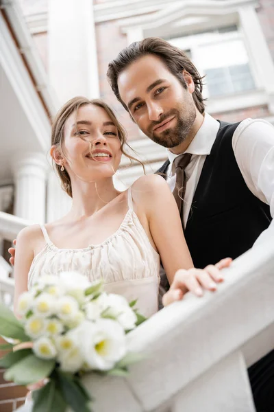 Vista de ángulo bajo de la joven feliz en vestido de novia y el novio en traje mirando a la cámara cerca de la casa - foto de stock
