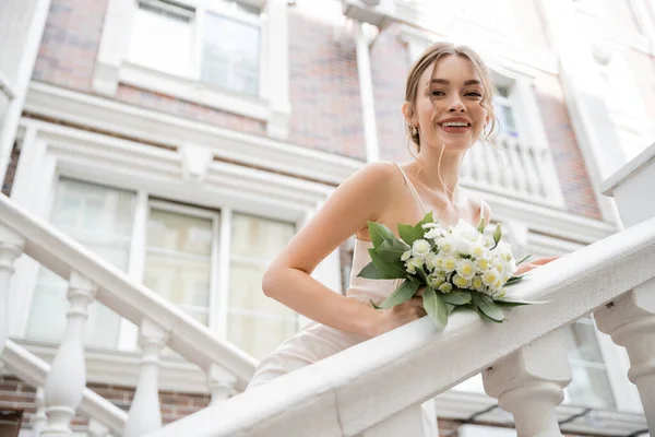 Vista de ángulo bajo de la novia feliz en vestido de novia con ramo de celebración y mirando a la cámara cerca de casa - foto de stock