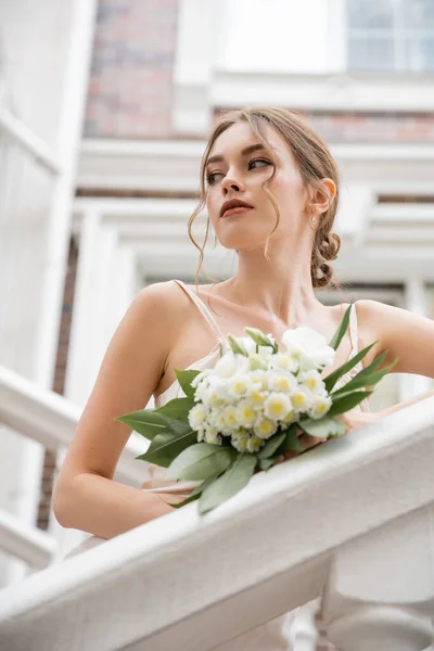 Low angle view of bride in wedding dress holding bouquet and looking away near house — Photo de stock