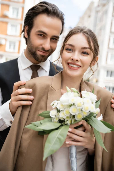 Retrato de novio feliz en chaleco abrazando novia alegre en blazer beige y celebración de ramo de boda - foto de stock