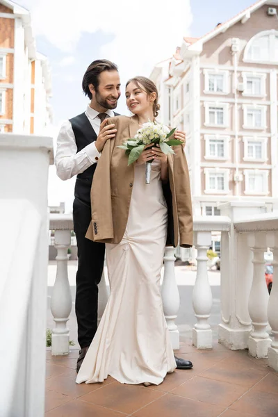 Longueur totale de marié heureux en gilet câlin mariée gaie en blazer beige et robe de mariée avec bouquet — Photo de stock