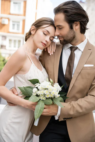 Young bride in wedding dress holding bouquet and leaning on groom in suit — Stockfoto