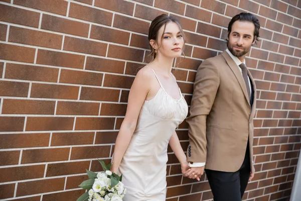 Young bride in wedding dress holding bouquet and hand of groom near brick wall — Stock Photo