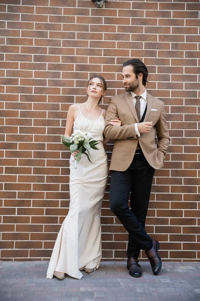 Full length of dreamy bride in wedding dress holding flowers near groom in suit while standing against brick wall — Stock Photo