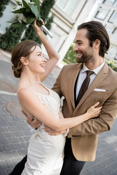 Novia alegre en vestido de novia celebración de ramo y sonriendo con el novio - foto de stock