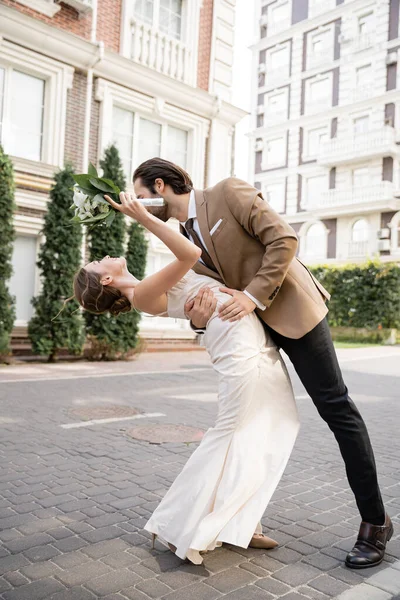 Comprimento total da noiva alegre no vestido de noiva segurando buquê e curvatura perto do noivo — Fotografia de Stock