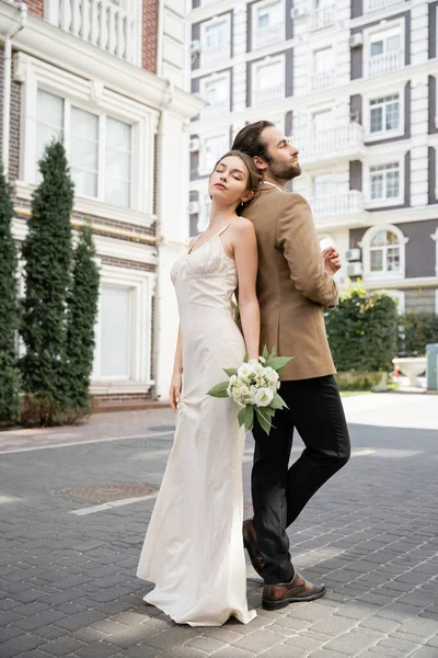 Повна довжина красивої нареченої у весільній сукні, що тримає букет і стоїть назад з нареченим — стокове фото