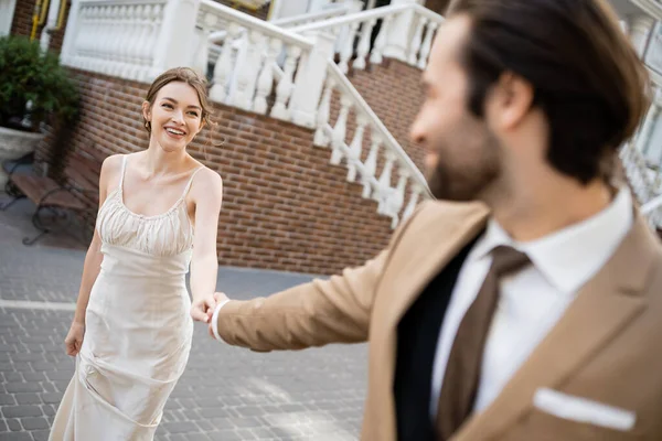 Blurred groom in suit holding hand of happy bride in white dress - foto de stock