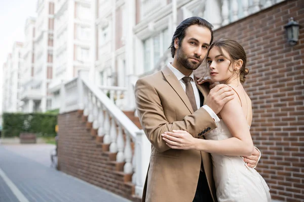 Groom in beige suit hugging sensual bride in wedding dress while standing on street — Stockfoto