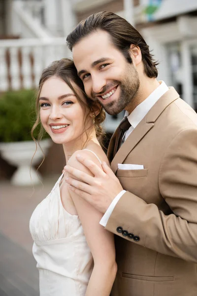 Happy groom in beige blazer hugging gorgeous bride in white wedding dress — Stock Photo
