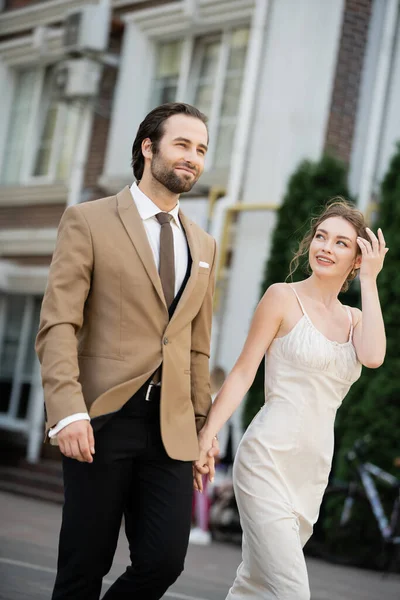 Happy bride in wedding dress looking at groom while holding hands on street — Stock Photo