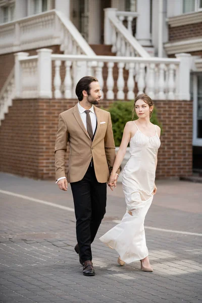 Full length of bride in wedding dress holding hands with groom while holding hands on street — Photo de stock