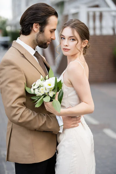 Bearded groom hugging bride in wedding dress holding blooming flowers — Foto stock