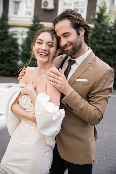 Portrait of positive and bearded man hugging happy bride in wedding dress — Stock Photo