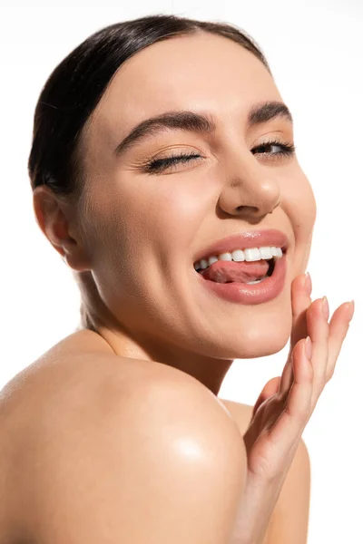 Позитивна жінка з натуральним макіяжем і голим плечем, торкаючись щоки, стирчить з язика ізольовано на білому — стокове фото