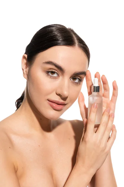 Brunette woman with bare shoulders looking at camera while holding bottle with moisturizing serum isolated on white — стоковое фото