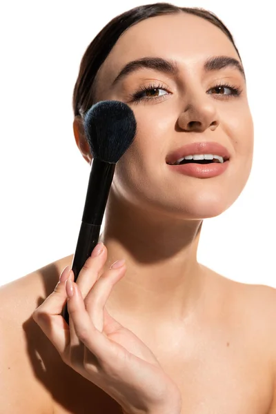 Brunette woman with bare shoulders holding soft powder brush near cheek isolated on white — Stock Photo