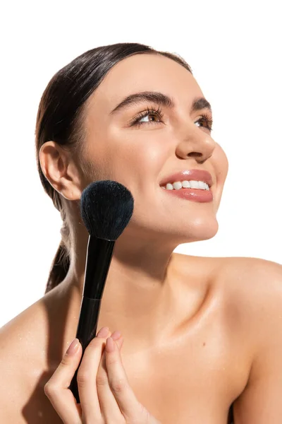 Mujer satisfecha con hombros desnudos sosteniendo un cepillo de polvo suave cerca de la mejilla aislada en blanco - foto de stock