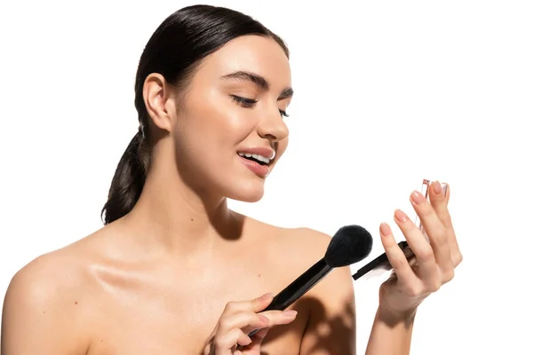 Cheerful woman with bare shoulders holding soft cosmetic brush near powder blush isolated on white — Fotografia de Stock