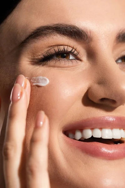 Close up view of joyful young woman applying cream on cheek — Stock Photo