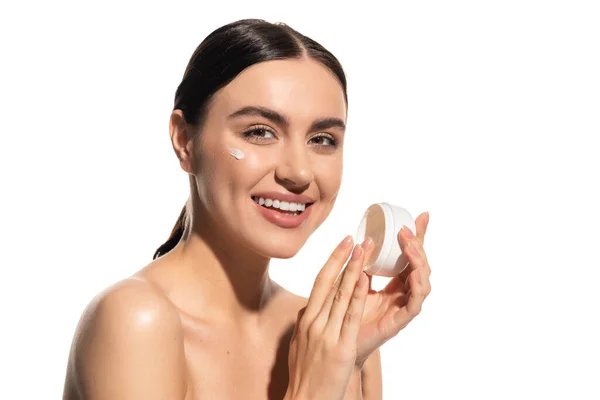Jovem mulher positiva com ombros nus segurando jar com creme hidratante no rosto isolado no branco — Fotografia de Stock