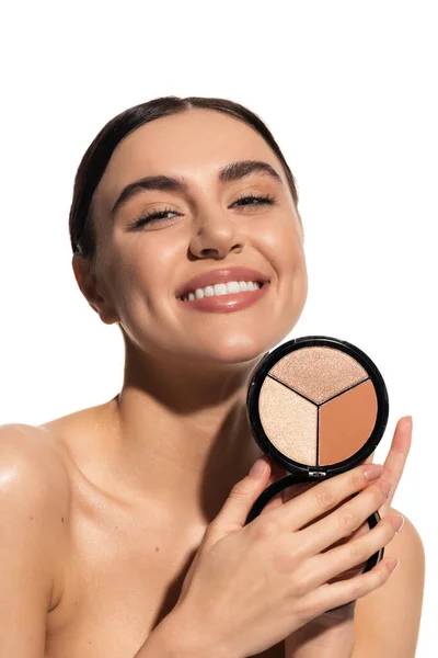 Positive young woman with bare shoulders holding bronzer and highlighter palette isolated on white — Stock Photo