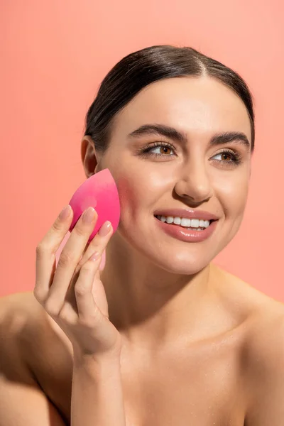 Happy young woman applying makeup foundation with beauty sponge isolated on pink — Stock Photo