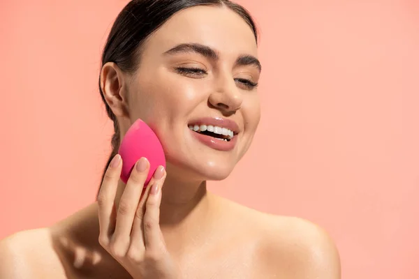 Happy woman applying face foundation with beauty sponge isolated on pink — Stock Photo