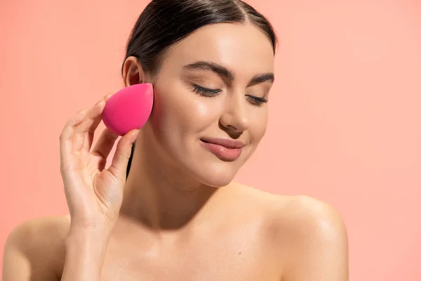 Pleased woman applying face foundation with makeup sponge isolated on pink — Stock Photo