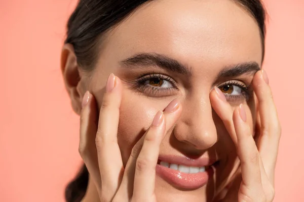 Close up view of cheerful young woman touching face while looking at camera isolated on pink — Stock Photo