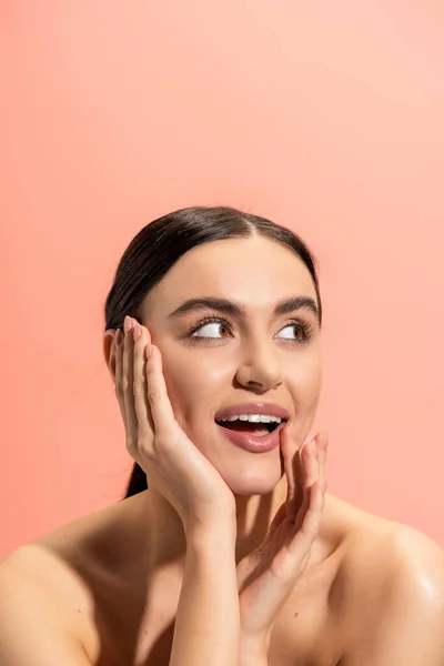 Jeune femme étonnée avec les épaules nues regardant loin tout en touchant la joue isolée sur rose — Photo de stock