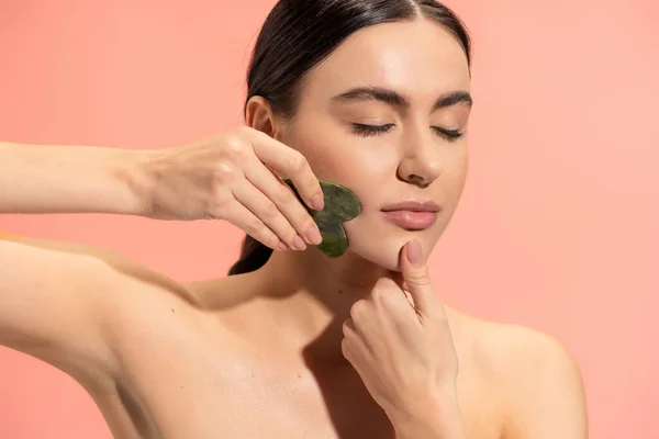 Mulher morena com pele macia fazendo massagem facial com raspador de jade isolado em rosa — Fotografia de Stock