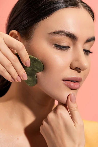 Close up view of brunette woman with soft skin doing face massage with jade scraper isolated on pink — Stock Photo