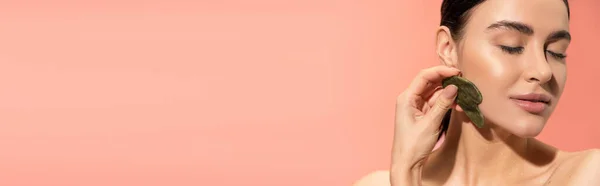Brunette femme à la peau douce faisant massage du visage avec grattoir en jade isolé sur rose, bannière — Photo de stock