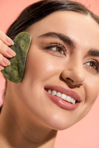 Close up de mulher feliz com pele macia fazendo massagem facial com raspador de jade isolado em rosa — Fotografia de Stock