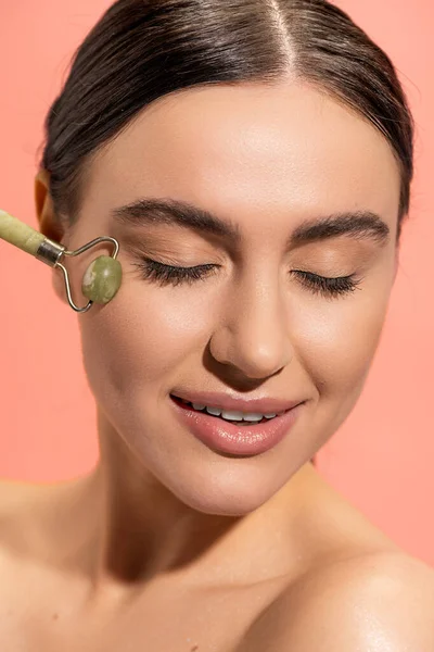 Cheerful young woman doing face massage with jade roller isolated on pink — Stock Photo